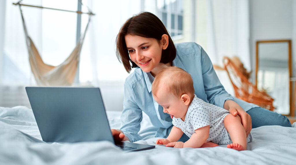 MamaGrow Beitragsbild - Eine Frau arbeitet während der Elternzeit, auf dem Bett an ihrem Laptop von zu Hause aus mit ihrem Kind neben an. Ein Symbolbild für Jobs für Mütter von zu Hause aus.