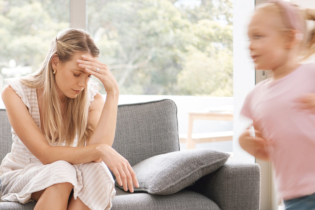 Überforderte Mutter Symptome - Auf einem Sofa sitzende Mutter ist ersichtlich überfordert und hat Kopfschmerzen, während Kind ihr in der Wohnung rennt