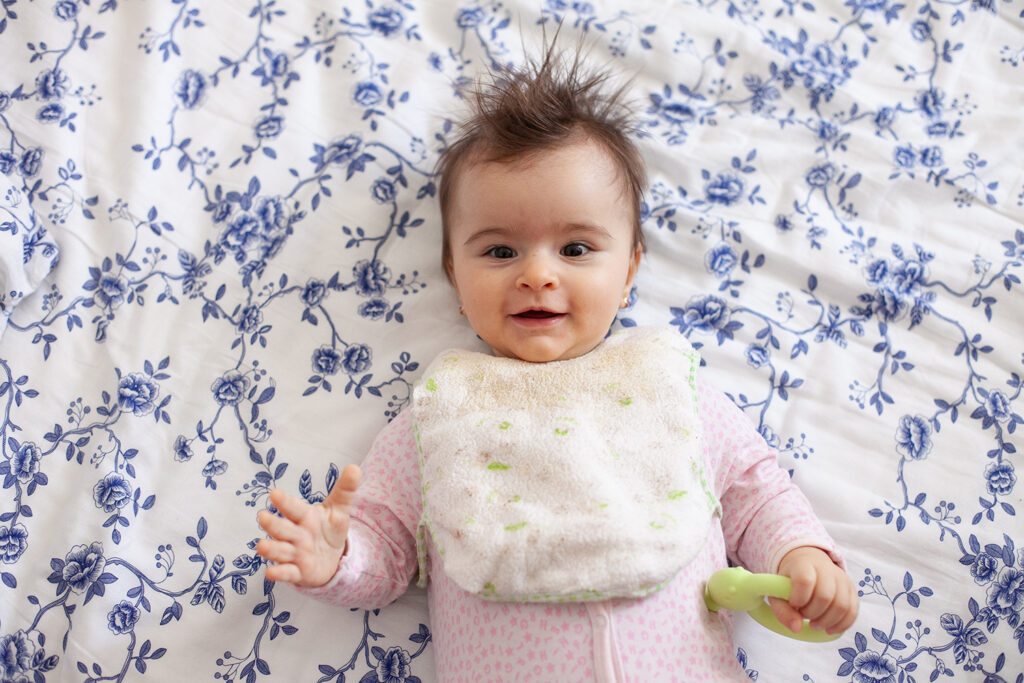 Babys erster Haarschnitt - Ein Baby mit zerzausten Haaren liegt auf dem Bett und lächelt in die Kamera.