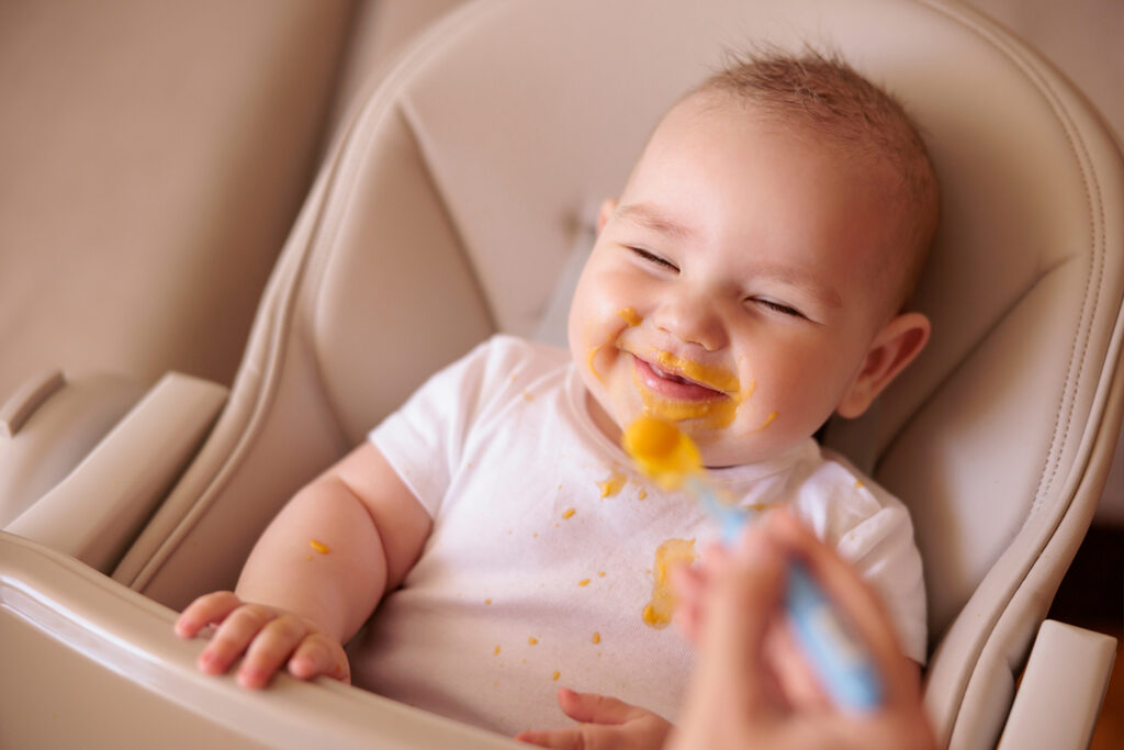 Nahaufnahme eines Babys, das von seiner Mutter mit Babybrei aus Kartoffeln gefüttert wird, welcher zuvor eingefroren wurde und das Baby nun um den Mund hat.