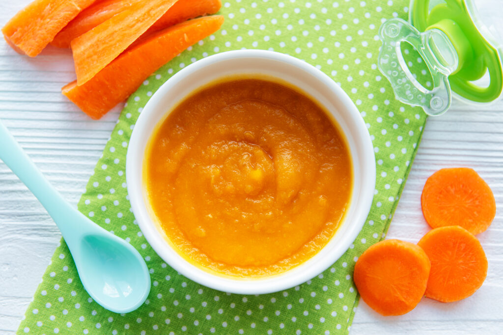 Rezepte für Babybrei ab dem 4., 5. und 6. Monat. Auf dem Tisch liegt eine Schüssel worin Babybrei aus Karotten und Kürbis enthalten ist.