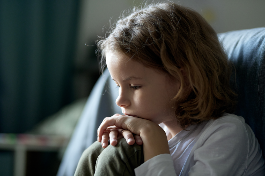Bipolare Störung bei Kindern - Nahaufnahme eines depressiven Kindes, dass nachdenklich im Zimmern sitzt und auf dem Boden schaut.