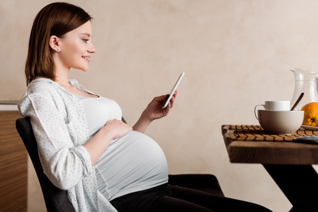 Eine Schwangere Frau sitzt am Frühstückstisch und surft auf ihrem Handy nach 'Frühstück für Schwangere', um Rezepte und Informationen darüber zu erhalten.