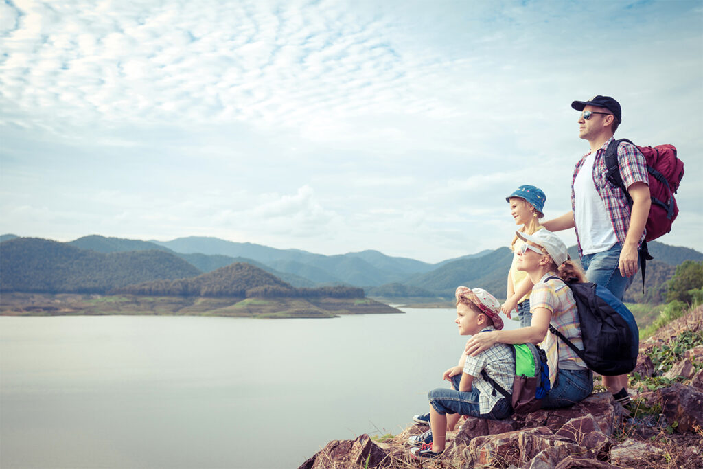 Urlaub mit Kindern am See - Eine Familie steht etwas erhöht und geniesst den Ausblick auf den See mit Bergpanorama im Hintergrund.
