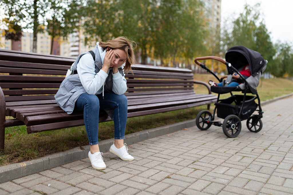 Depression Mutter - Eine Mutter mit Depressionen sitzt alleine auf einer Parkbank. Neben ihr befindet sich ihr Kinderwagen mit ihrem Baby. Symbolisch liegt ein grosser Abstand zwischen ihr und ihrem Baby.