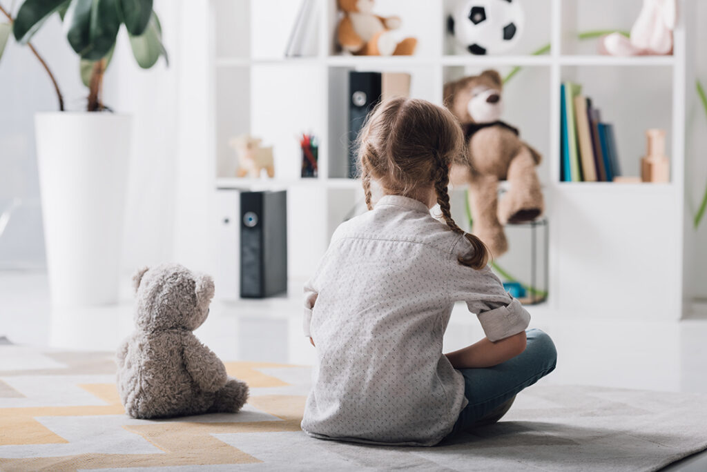 Kind wird ausgeschlossen - Rückansicht eines einsamen kleinen Kindes, das im Kinderzimmer mit Teddybär auf dem Boden sitzt.