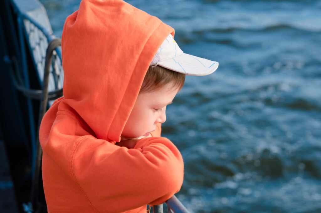 Reisekrankheit Kinder - Ein junge in einem orangen Hoodie und einer weissen Schirmmütze, schaut vom Boot aus aufs Wasser herab, weil ihm Unwohl ist. Ein Symbolbild für die Reisekrankheit bei Kindern.