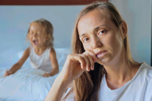 Herausforderung Wutanfall Kleinkind: Wann sind Wutanfälle bei Kindern nicht mehr normal? Porträt einer verärgerten, müden Mutter mit einem wütenden kleinen Mädchen im Hintergrund, das schreit und auf ein Kissen einschlägt.