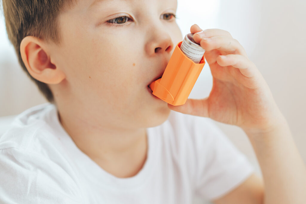 Was äussert sich Asthma bei Kindern? Nahaufnahme eines kleinen Jungen, der einen orangen Asthma-Inhalator verwenndet. Der Junge trägt ein weisses T-Shirt.