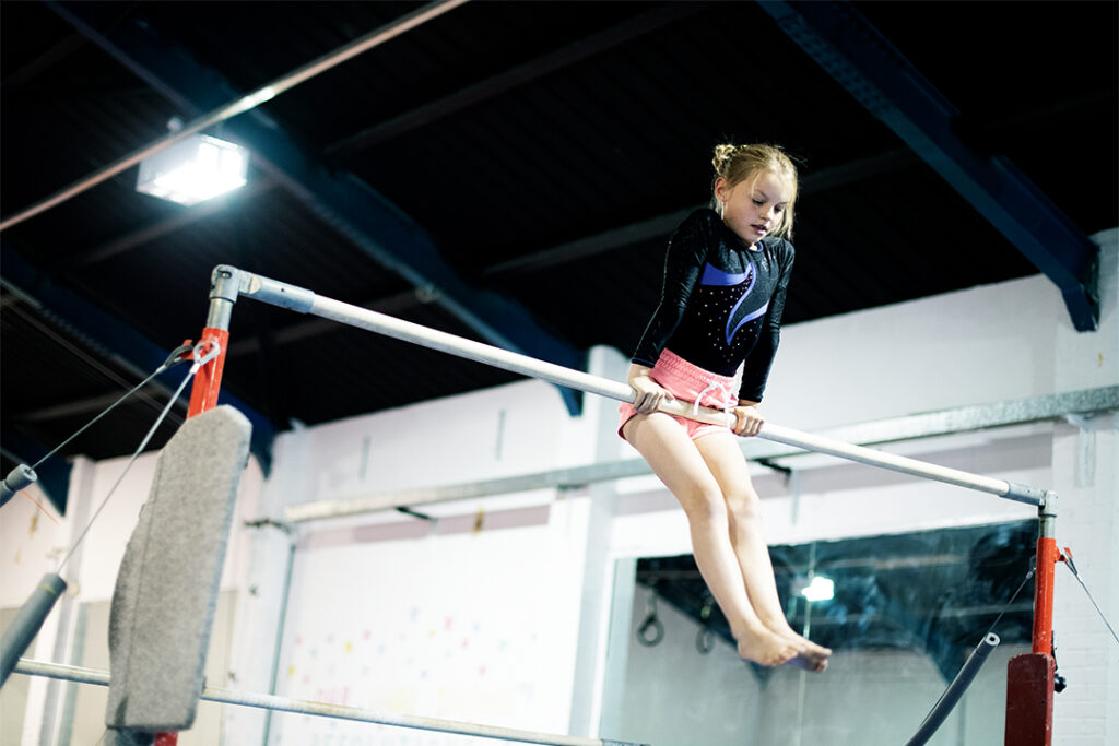 Geräteturnen für Kinder - Ein Mädchen turnt an der Reckstange in der Sporthalle und trägt ein Gymnastik Kostüm.