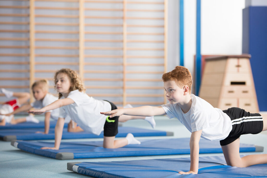 Bodenturnen für Kinder - Kinder in einer Turnhalle machen Bodenturnübungen auf blauen Matten. Im Hintergrund ist eine Sprossenwand zu sehen.