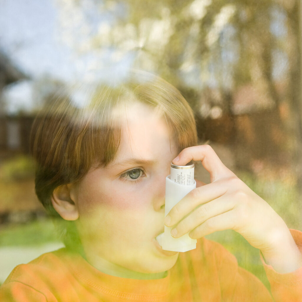 Ein Junge mit Asthma steht hinter dem Fenster und schaut raus. Er verwendet seinen Asthma-Inhalator, weil ihm die Pollen-Allergie zu schaffen macht. In der Spiegelung des Fensters ist ein blühender Baum zu sehen.