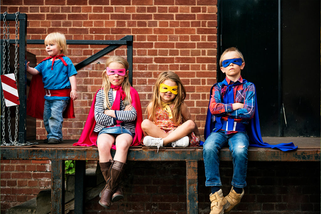 Wie stärke ich das Selbstbewusstsein meines Kindes? Kinder in Superhelden-Kostümen sitzen vor einer Zielgelmauer, als Symbolbild für Selbstbewusstsein bei Kindern.