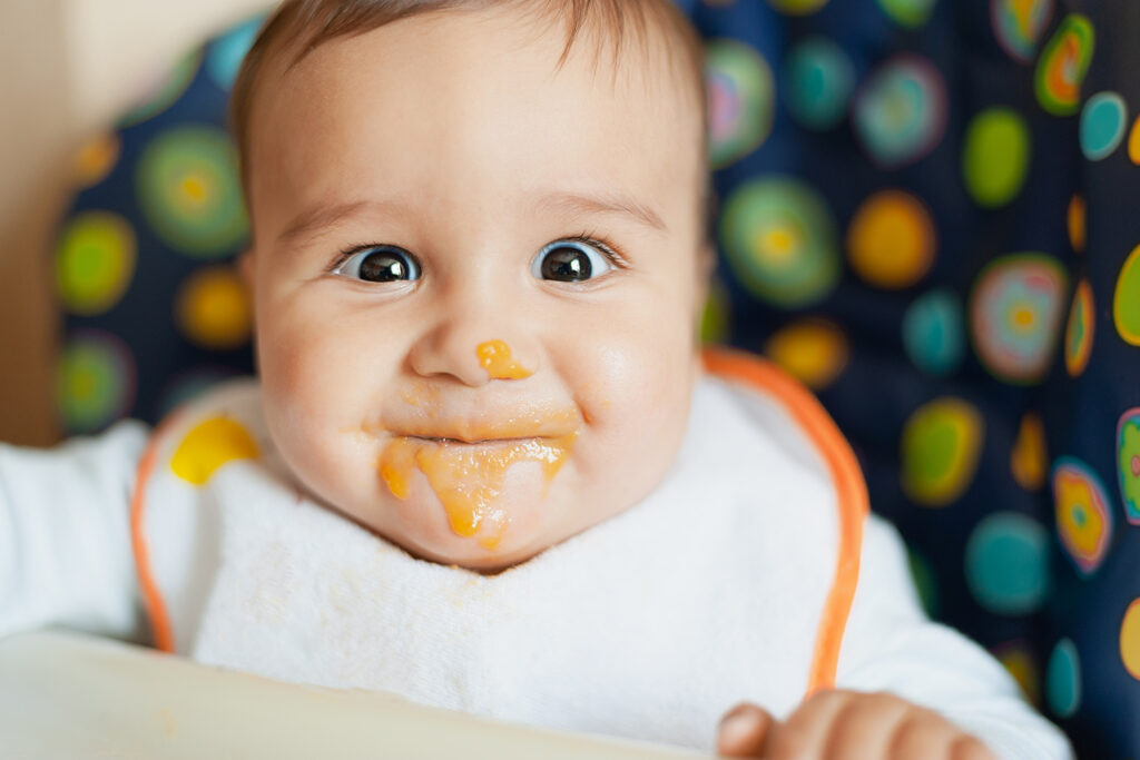 Nahaufnahme eines Babys das lustig reinschaut, nachdem es Babybrei gegessen hat und nun ein verschmiertes Gesicht hat.
